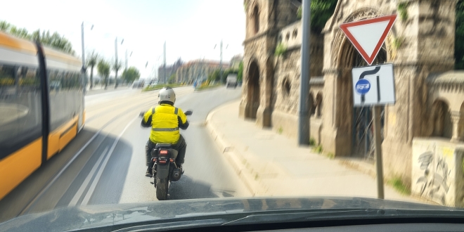 Autóból nézve egy motoros halad a budai rakparton mellette egy villamossal, mellette egy elsőbbségadás kötelező tábla.